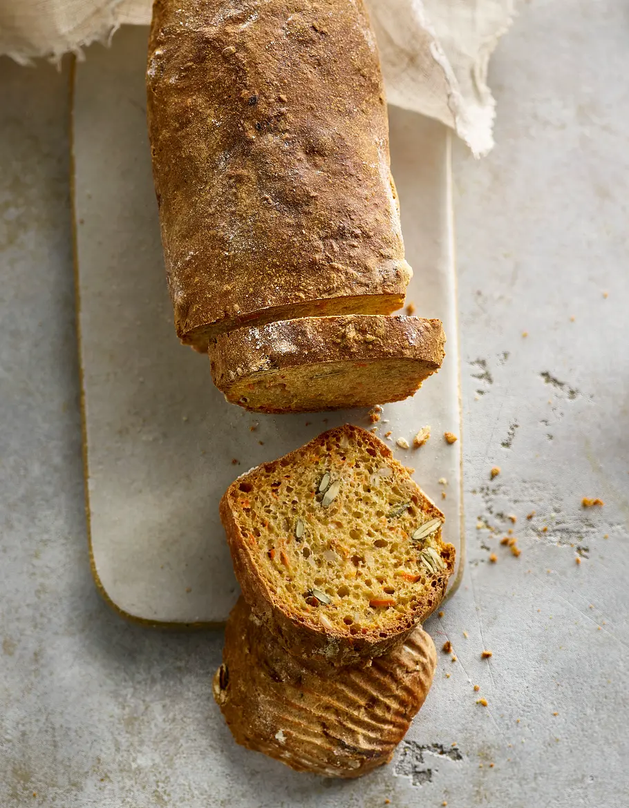 Krustenbrot aus dem Airfryer mit Dinkel und Karotte Das Krustenbrot mit Dinkelmehl und Karotte liegt auf einem Brettchen, zwei Scheiben sind abgeschnitten.