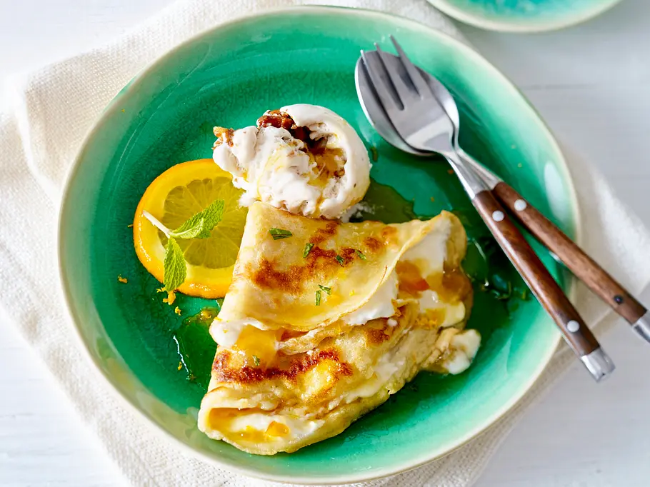 Crêpe à l’orange Rezept Zarter Crêpe, gefüllt mit Orangencreme, serviert mit einer Orangenscheibe und einer Kugel Eis auf einem grünen Teller.