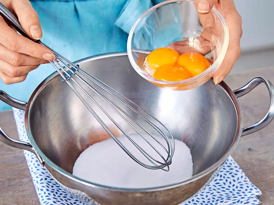 Erdbeer-Pistazien-Parfait Eine Schüssel mit Zucker, dazu eine Hand, die Eigelb aus einer kleinen Glasschale hineingibt, daneben ein Schneebesen, alles auf einem gemusterten Tuch.
