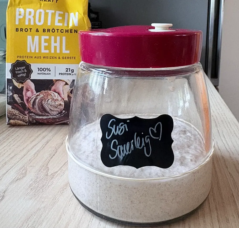 Leaven Mischen Ein Glas mit Sauerteig, rotem Deckel und der Aufschrift „Susi Sauerteig“ steht vor einer gelben Packung Proteinmehl.