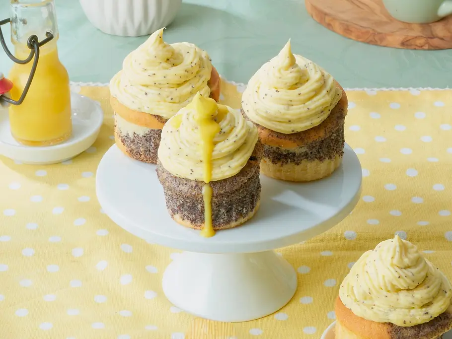 Vier Cupcakes mit gelbem Eierlikör-Creme-Topping stehen auf einer Etagere und einem gelben Tuch, daneben ein Glas Eierlikör.