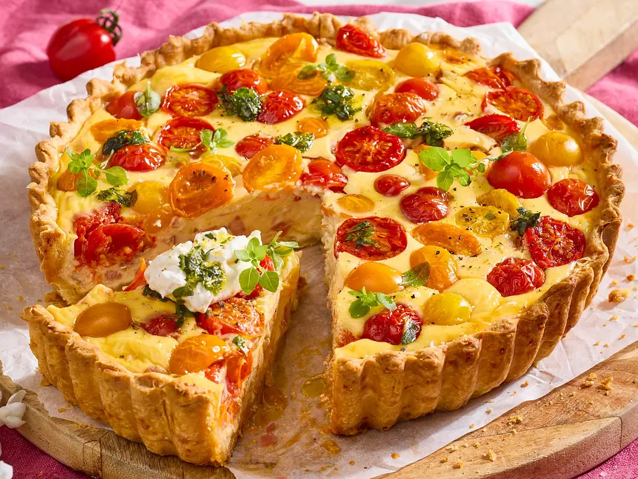 Tomaten-Quiche mit gelben und roten Kirschtomaten, Frischkäse und Burrata, auf rundem Holzbrett und Backpapier serviert.