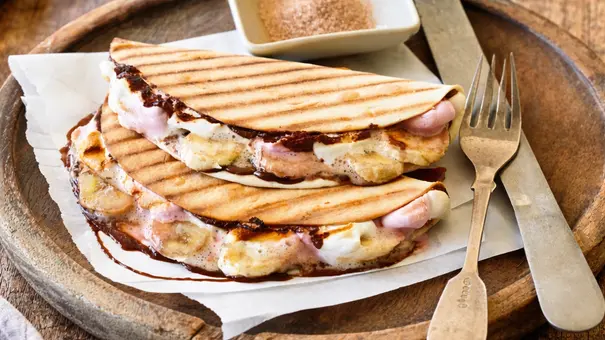 Zwei goldbraun gegrillte Tacos mit sichtbaren Grillstreifen, gefüllt mit geschmolzenen Marshmallows, Bananenscheiben und Schokoladensoße. Auf Backpapier in einer runden Holzschale angerichtet, daneben eine kleine Schale mit Zimtzucker sowie Messer und Gabel. - Foto: Image Professionals