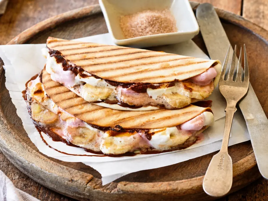 Zwei goldbraun gegrillte Tacos mit sichtbaren Grillstreifen, gefüllt mit geschmolzenen Marshmallows, Bananenscheiben und Schokoladensoße. Auf Backpapier in einer runden Holzschale angerichtet, daneben eine kleine Schale mit Zimtzucker sowie Messer und Gabel.