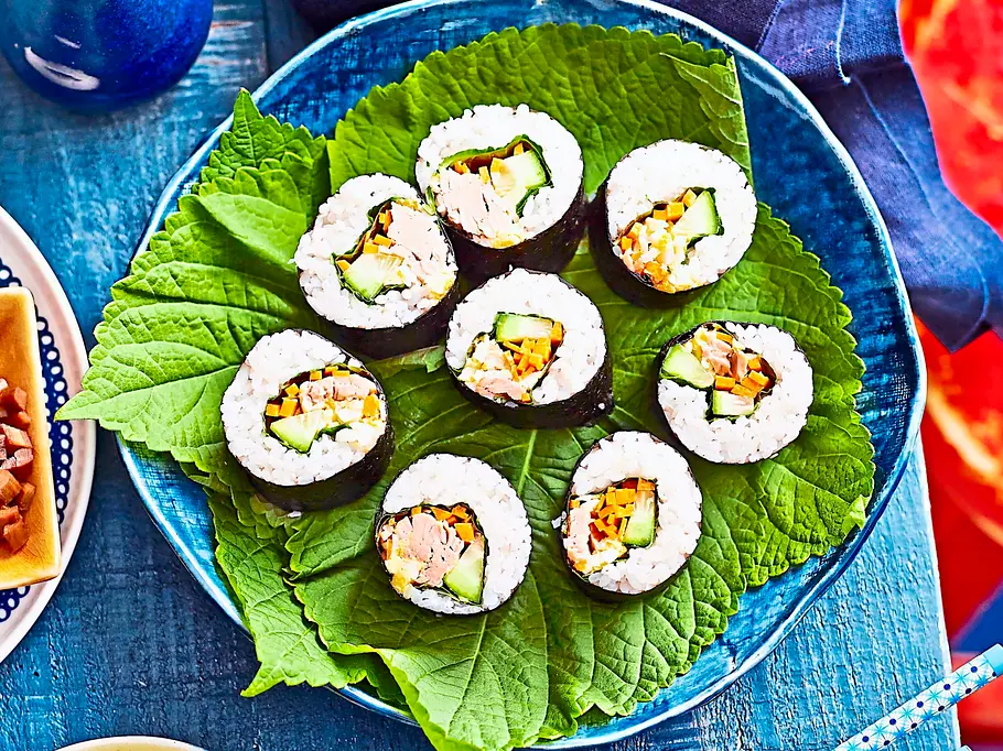Tuna Kimbap Tuna Kimbap-Rollen, gefüllt mit Thunfisch, Gurke und Karotte, angerichtet auf grünen Blättern auf einem blauen Teller.