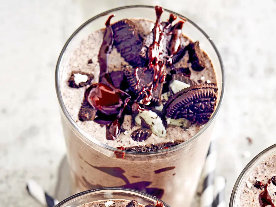 Drei Gläser cremiger Cookie & Cream Milchshake, getoppt mit zerbröselten Schokokeksen und Schokosauce, dazu gestreifte Papierstrohhalme auf einer hellen Unterlage.