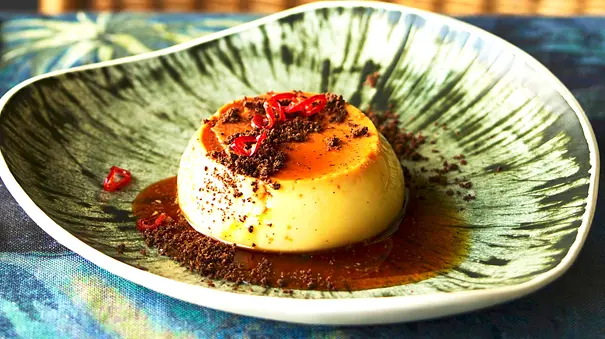 Cremiger Flan mit goldener Karamellschicht, garniert mit dunkler Schokolade und feiner Chilischeibe, serviert auf einem künstlerisch gemusterten Teller. - Foto: LECKER @ Bauer Media Group