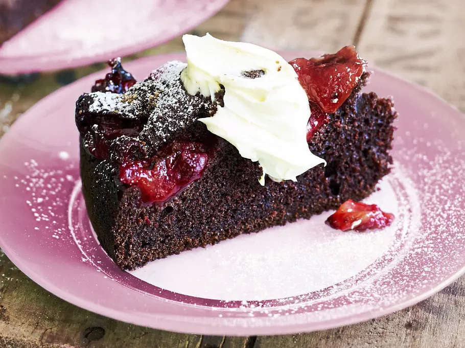 Ein saftiges Stück Schoko-Espresso-Kuchen liegt auf einem rosa Teller, belegt mit roten Fruchtstücken und einem Klecks Sahne. Puderzucker ist darüber gestäubt, im Hintergrund steht eine Metallkanne.