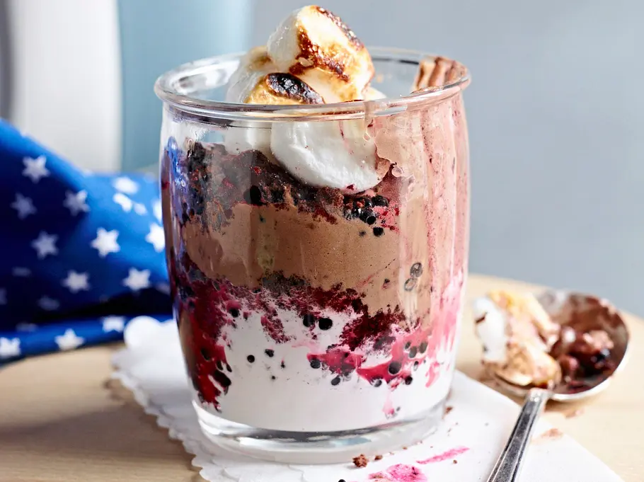 Schichtdessert im Glas mit cremiger Schokoladenmousse, Beeren, gerösteten Marshmallows und Sahne auf einem kleinen Holzbrett, daneben ein Löffel.