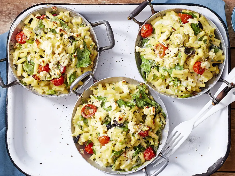 Cremiges Mac-&-Cheese-Gratin mit zart überbackener Käseschicht, goldgelben Makkaroni, saftigen Kirschtomaten und frischen grünen Blättern, angerichtet in drei kleinen runden Metallpfannen auf einem weißen Tablett aus Emaille.