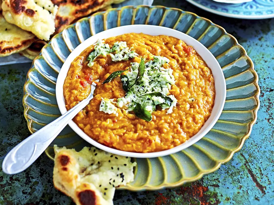 Linsen-Dal mit Kokos-Chutney Eine Schale mit orangefarbenem Linsengericht und grün-weißer Garnitur steht neben Fladenbroten und einer Schale grünem Dip.