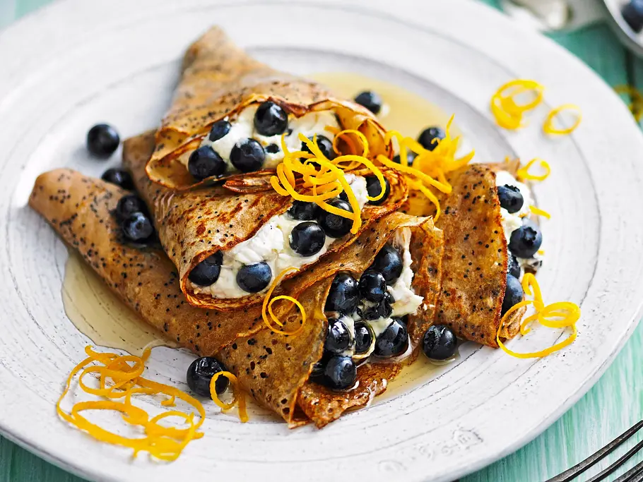 Mohn-Crêpes mit Mascarpone Gefüllte Mohn-Crêpes mit Mascarpone und frischen Blaubeeren, garniert mit feinen Orangenzesten und Ahornsirup, angerichtet auf einem rustikalen weißen Teller auf grün-blauem Holztisch.