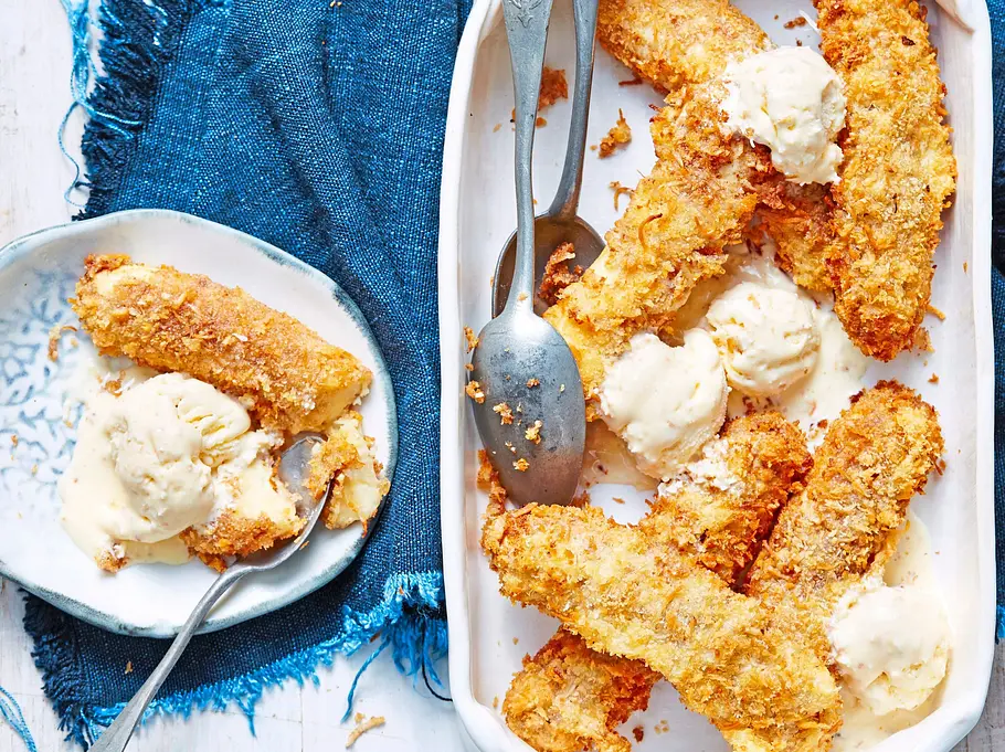 Knusprig frittierte Bananen in Kokos-Panade, serviert mit cremigen Kugeln Vanilleeis in einer weißen Auflaufform auf blauem Stoff. Daneben ein kleiner Teller mit einem Stück Banane und Eiscreme.