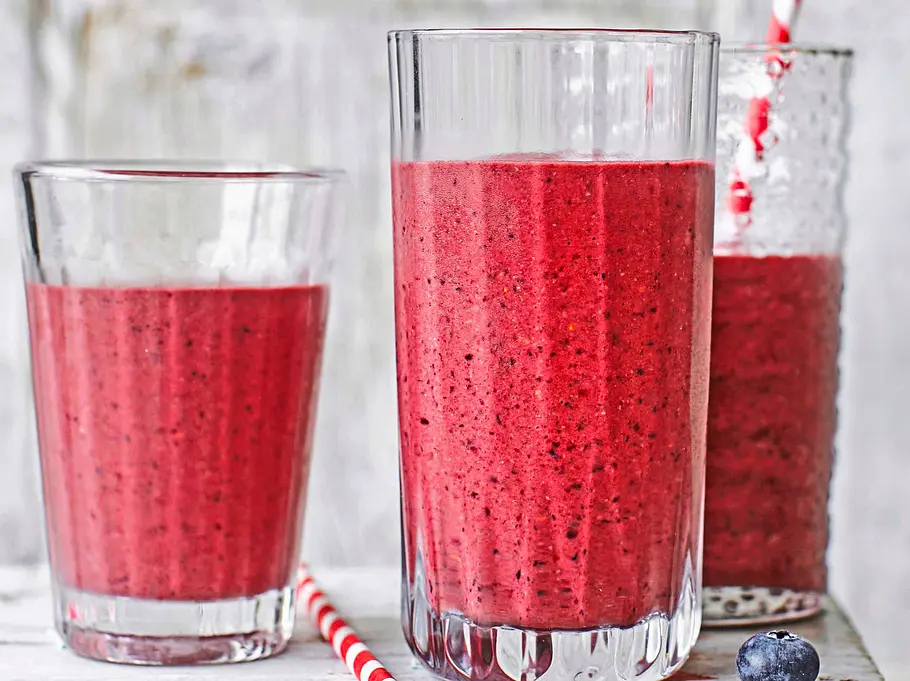 Drei Gläser mit violettem Heidelbeer-Quark-Shake auf einem rustikalen Holzbrett, daneben ein kleines Glas mit frischen Heidelbeeren und zwei gestreifte Trinkhalme.