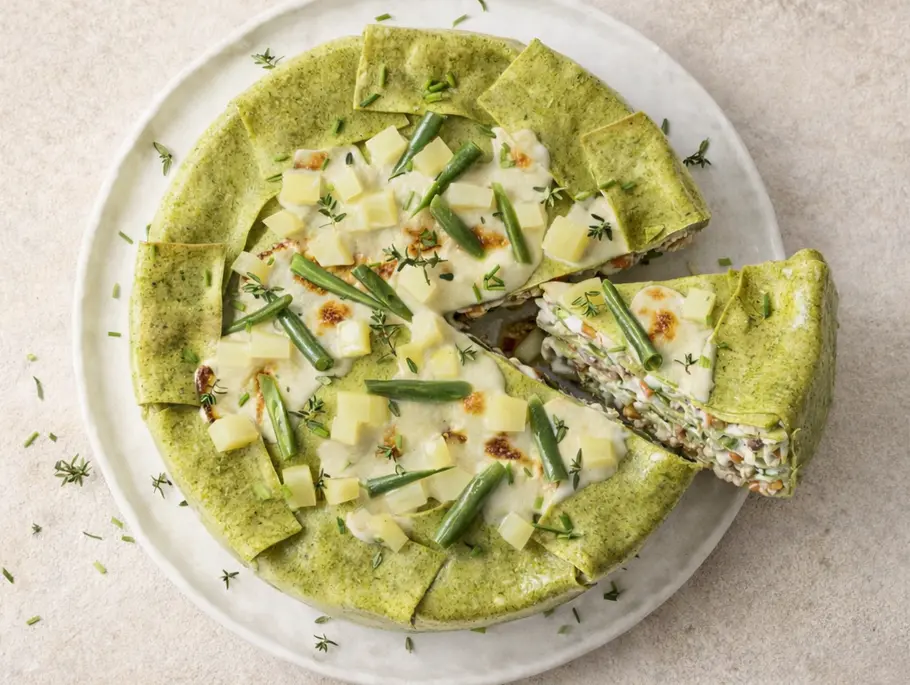 Runde Lasagne mit grünen Nudelplatten, gefüllt mit Hähnchen, grünen Bohnen und weißen Bohnen, cremig überbacken und mit frischen Kräutern bestreut, serviert auf einem weißen Teller, ein Stück herausgeschnitten.