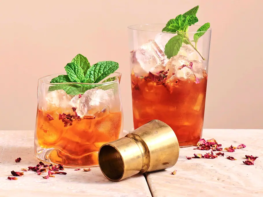 Zwei Gläser mit orientalischem Tea Punch auf Eis, garniert mit frischer Minze und Rosenblüten, serviert auf einer hellen Marmorplatte mit goldenem Messbecher.