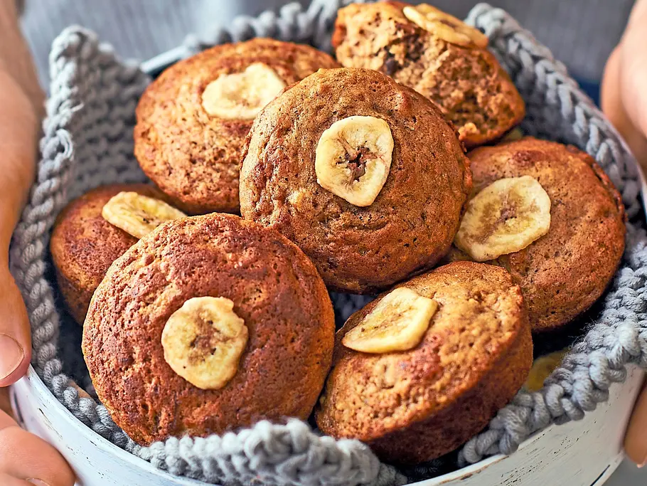 Kleine Bananenbrot-Mini-Pies mit goldbrauner Kruste und getrockneter Bananenscheibe auf jedem Gebäck, angerichtet in einer weißen Schale auf einem gestrickten, grauen Untersetzer.