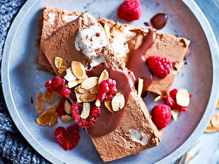 Cremiger Schokomousse-Eiskuchen in Scheiben, verführerisch garniert mit frischen Himbeeren, gerösteten Mandeln und einem Klecks Schokoladensauce, serviert auf einem modernen, hellen Teller mit Vintage-Besteck.