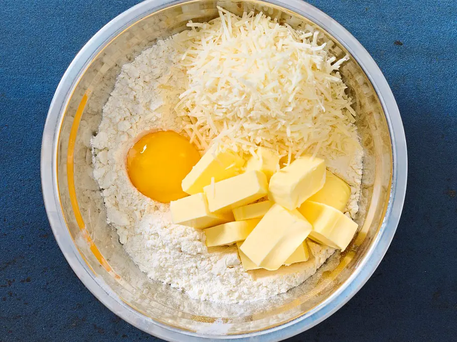 Eine Edelstahlschüssel mit Mehl, geriebenem Gouda, einem Eigelb und mehreren Stücken Butter auf einer blauen, strukturierten Unterlage.