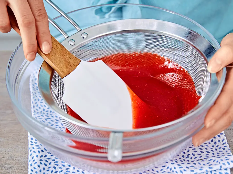 Erdbeer-Pistazien-Parfait Ein leuchtend rotes Erdbeerpüree wird mit einem Spatel durch ein Sieb in eine Glasschüssel gestrichen, die auf einem gemusterten Tuch steht.