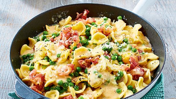Pasta in cremiger Carbonara-Soße mit Erbsen und Schinkenstücken in einer Pfanne auf grünem Untersetzer. - Foto: House of Food / Bauer Food Experts KG