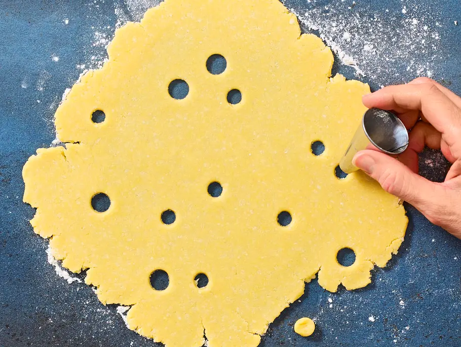 Ausgerollter gelber Teig auf bemehlter, blauer Unterlage, aus dem mit einer kleinen runden Ausstechform Löcher ausgestochen werden.