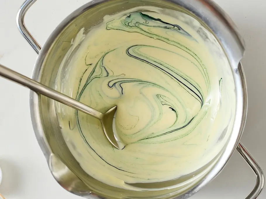 Geschmolzene weiße Kuvertüre im Topf, mit grüner Speisefarbe leicht marmoriert, daneben ein kleines Schälchen mit grüner Lebensmittelfarbe und Holzstäbchen auf heller Arbeitsfläche.