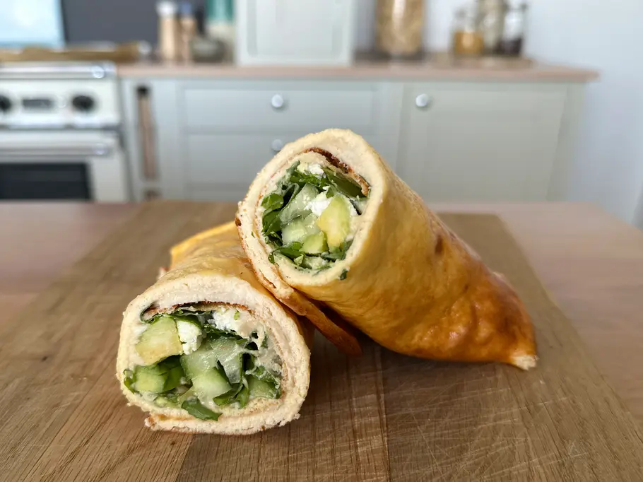 2-Zutaten-Protein-Wrap Aufgeschnittener Wrap mit cremiger Frischkäsefüllung, Avocado, Gurke und Salat auf einem Holzbrett in einer Küche.