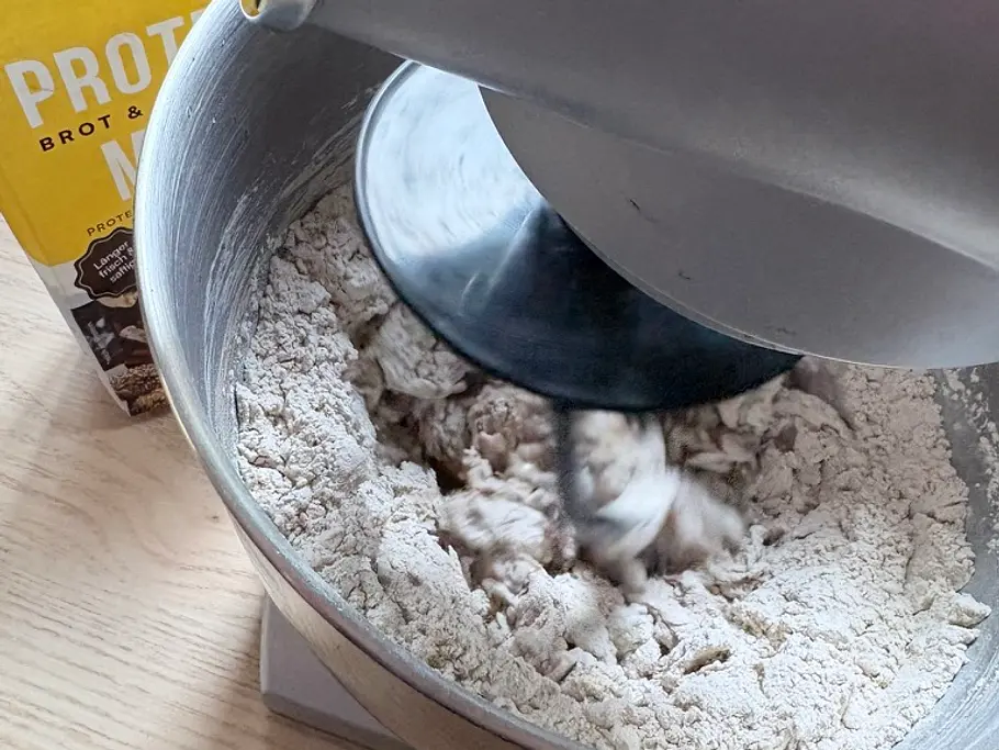 Protein-Sauerteigbrot Schritt Autolyse Eine Küchenmaschine knetet Teig mit Mehl in einer Metallschüssel. Daneben steht eine gelbe Packung Proteinmehl.