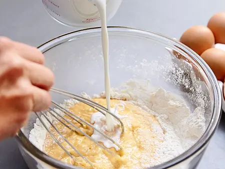 Rührende Teig-Melange Milch wird langsam zu einer Mehl-Ei-Mischung in einer Glasschüssel gegossen und mit einem Schneebesen verrührt, daneben liegen Eier und Eierschalen.