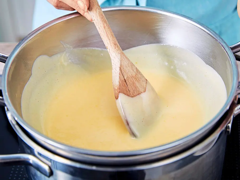 Erdbeer-Pistazien-Parfait Eine helle Creme wird in einem Topf mit einem Holzlöffel auf dem Herd gerührt, der Topf steht auf einem Herdfeld.