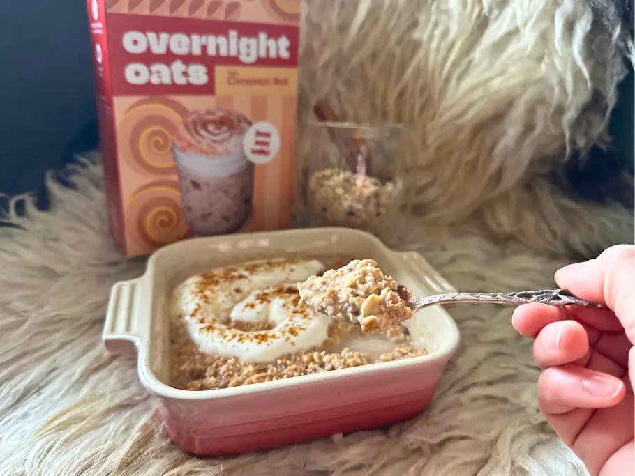 Eine Schale mit 3Bears Overnight Oats Typ Cinnamon Roll samt Joghurt-Swirl als Topping wird von einer Hand angelöffelt. Im Hintergrund ist die Packung sichtbar.