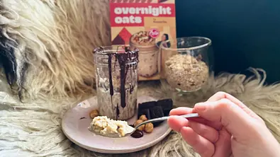 Eine Hand hält einen Löffel der zubereiteten Nussecken-Overnight-Oats von 3Bears x Sallys vor die Kamera. Dahinter ist die Packung, die abgefüllten Haferflocken-Mischung und die mit Schokolade garnierte Portion zu sehen - Foto: LECKER @ Bauer Media Group / Nicole Molitor
