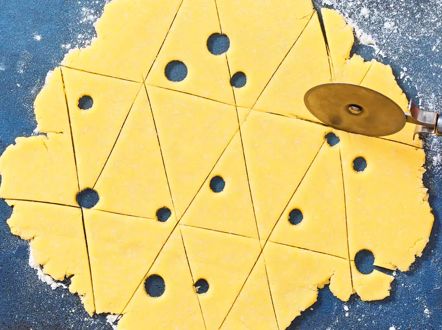 Der ausgerollte, gelbe Teig mit ausgestochenen Löchern wird mit einem Pizzaschneider in dreieckige Stücke geschnitten, liegt dabei auf bemehlter, blauer Unterlage.