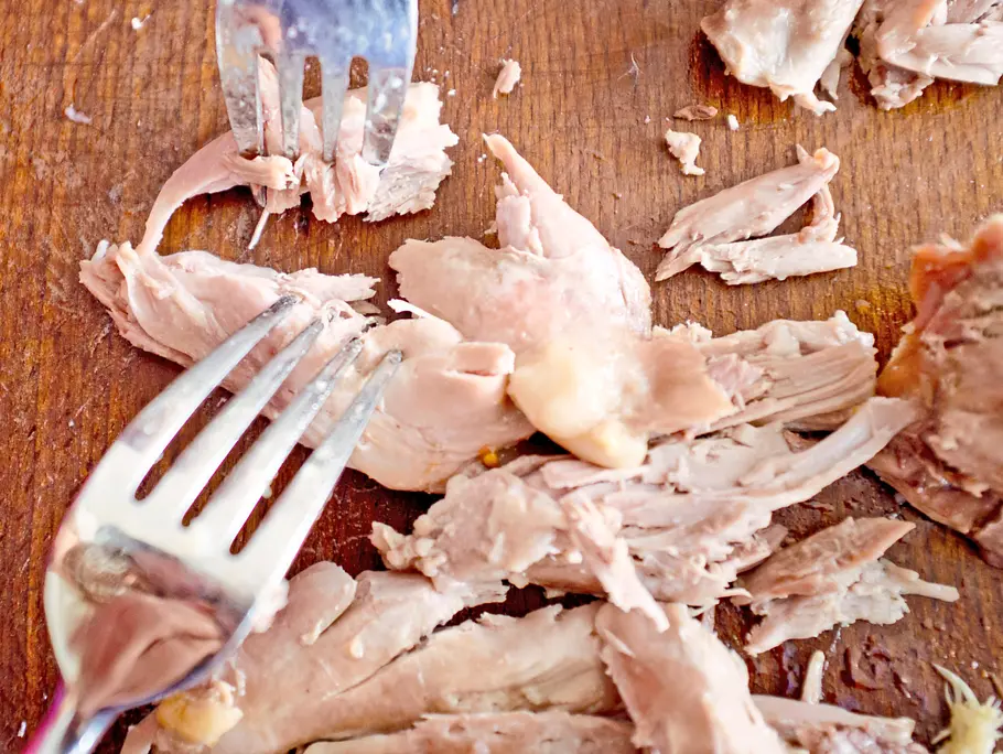 Mit zwei Gabeln wird gegartes Geflügelfleisch auf einem Holzbrett grob auseinander gezupft.