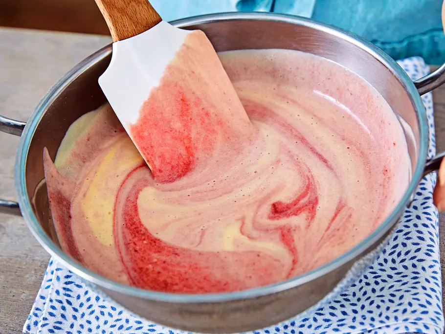 Erdbeer-Pistazien-Parfait Erdbeerpüree wird mit einem Spatel unter eine helle Creme in einer Metallschüssel auf einem gemusterten Tuch gemischt, es entstehen rot-gelbliche Wirbel.