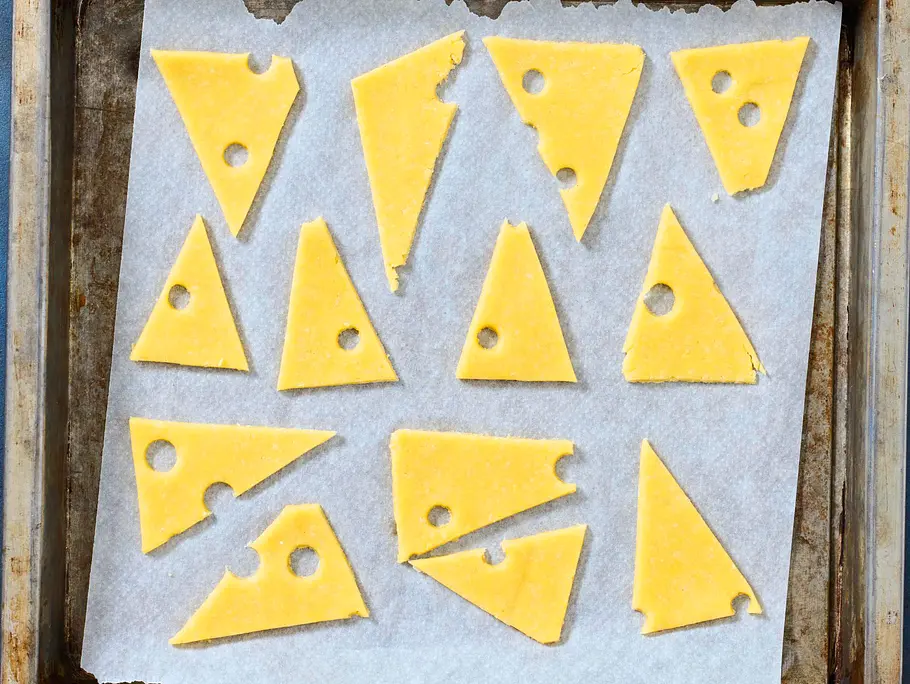 Dreieckige, gelbe Teigstücke mit ausgestochenen Löchern liegen auf Backpapier in einem Backblech, bereit zum Backen.