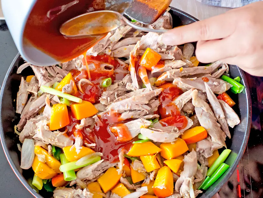 Gezupftes Fleisch, Kürbiswürfel, Frühlingszwiebeln und Schalotten werden in einer Pfanne angebraten, Ketchupsoße wird dazugegossen.