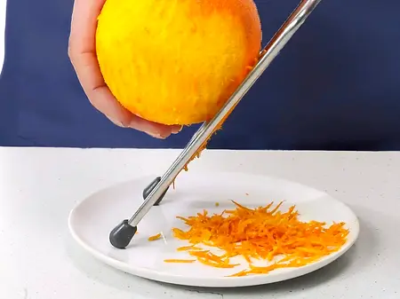 Mehr Orange! Mit einer feinen Reibe wird Orangenschale abgerieben und auf einem kleinen weißen Teller gesammelt.