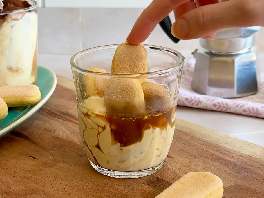 5 Minuten Tiramisu Rezept Ein Glas mit Espresso und Mascarpone-Creme. Eine Hand drückt halbe Löffelbiskuits in die Creme