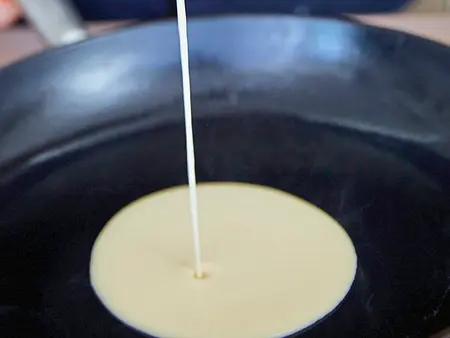 Crêperie zu Hause Flüssiger Crêpeteig wird mit einer Kelle in eine heiße, geölte Pfanne gegossen.