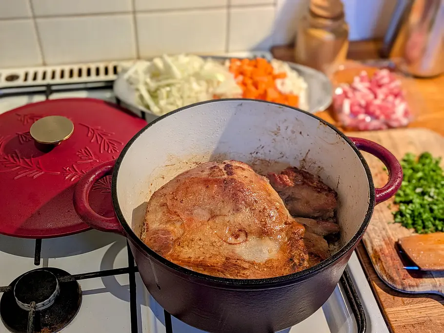 Die Lammkeule liegt in einem roten Bräter und ist goldbraun angebraten, daneben Gemüse, Speck und gehackte Kräuter.