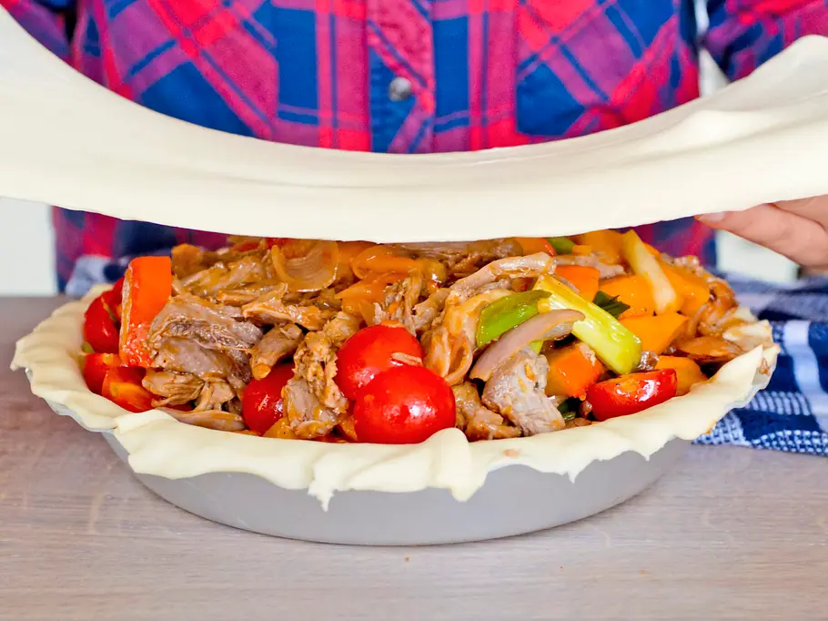 Füllung aus Fleisch und Gemüse in einer Teigform, wird mit einer zweiten Schicht Blätterteig bedeckt, die an den Rändern leicht verdreht wird.
