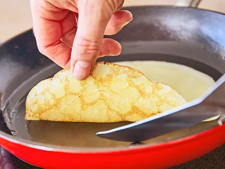 Bitte wenden Ein Crêpe wird in einer Pfanne vorsichtig mit der Hand und einem Pfannenwender gewendet.
