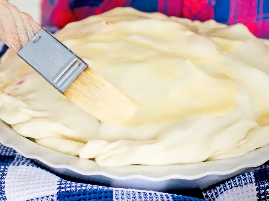 Die mit Teig bedeckte Pie wird mit einer Eier-Milch-Mischung bepinselt, auf einer Backunterlage vorbereitet.