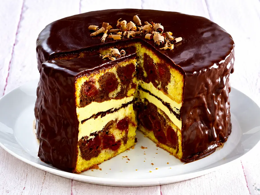 Doppelte Donauwelle Eine angeschnittene Torte mit Schokoglasur, marmorierten Böden, Kirschen und hellen Cremeschichten steht auf einer weißen Platte.