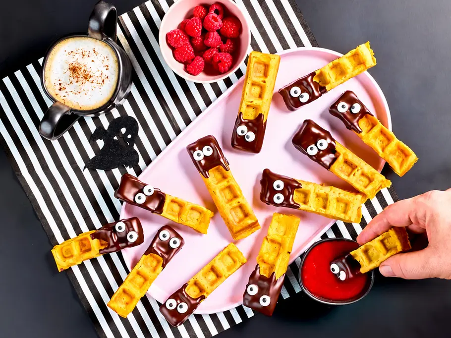 Monster-Waffel-Pommes Waffelstäbchen mit Schokolade und Zuckeraugen liegen auf einer rosa Platte. Daneben stehen Himbeeren, Fruchtsauce und Cappuccino.