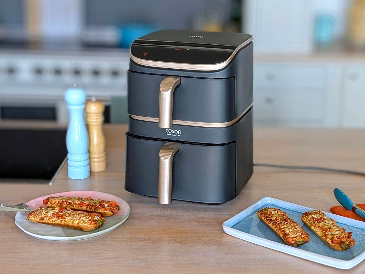 Der Cosori Turbo Tower Pro Smart steht auf einer Arbeitsfläche. Daneben eine Salz- und eine Pfeffermühle. Im Vordergrund zwei Teller mit jeweils zwei Pizzabaguettes.