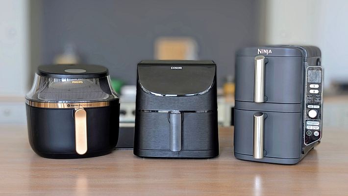 Auf einer Arbeitsfläche aus Holz stehen drei Airfryer nebeneinander: der Philips 3000 Series 6,2L, der Cosori 5,5L XXL und der Ninja Double Stack XL. - Foto: Constanze Brinkschulte / LECKER.de