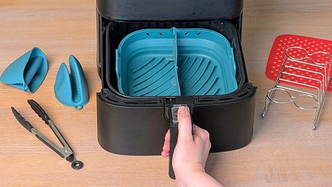 Auf einer Tischplatte aus Holz steht eine Heißluftfritteuse, deren Schublade von einer Hand herausgezogen wird. In der Schublade liegt eine türkise Silikonform. Rundherum liegt diverses anderes Airfryer-Zubehör. - Foto: LECKER.de/Constanze Brinkschulte